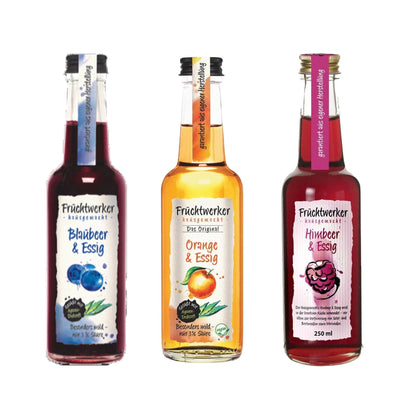 Fruchtwerker Essig-Set mit Flaschen für Blaubeere, Orange und Himbeere auf holzfarbigem Hintergrund.